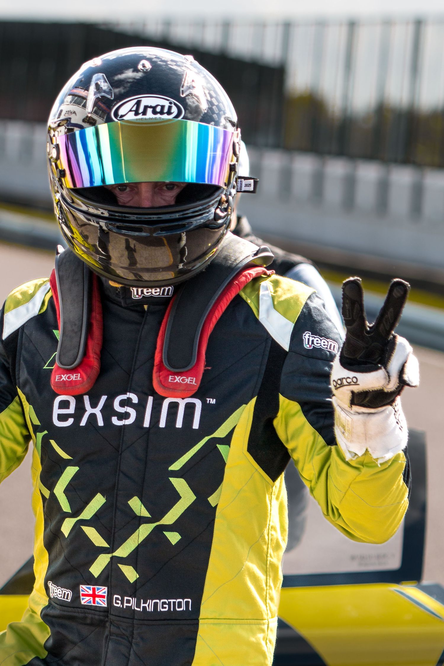 Racing driver in exsim-sponsored yellow and black race suit and Arai helmet giving a peace sign victory gesture.