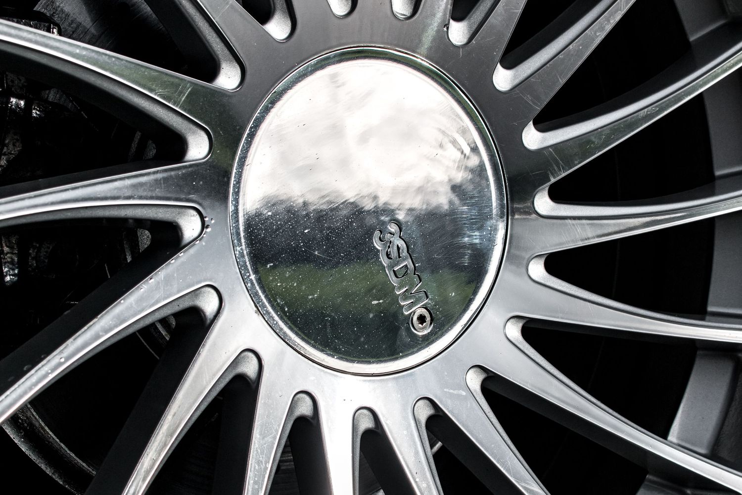 Close-up of a chrome GMC alloy wheel hub cap with multi-spoke rim design, reflecting clouds and trees.