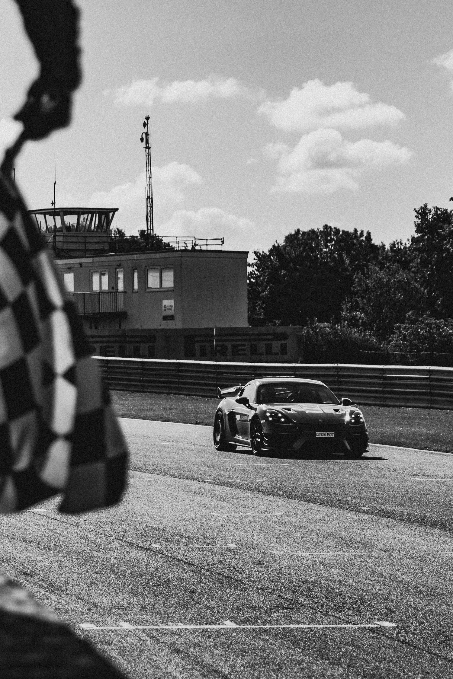 Black and white photo of a sports car on a racing circuit with a checkered flag in the foreground and control tower behind.