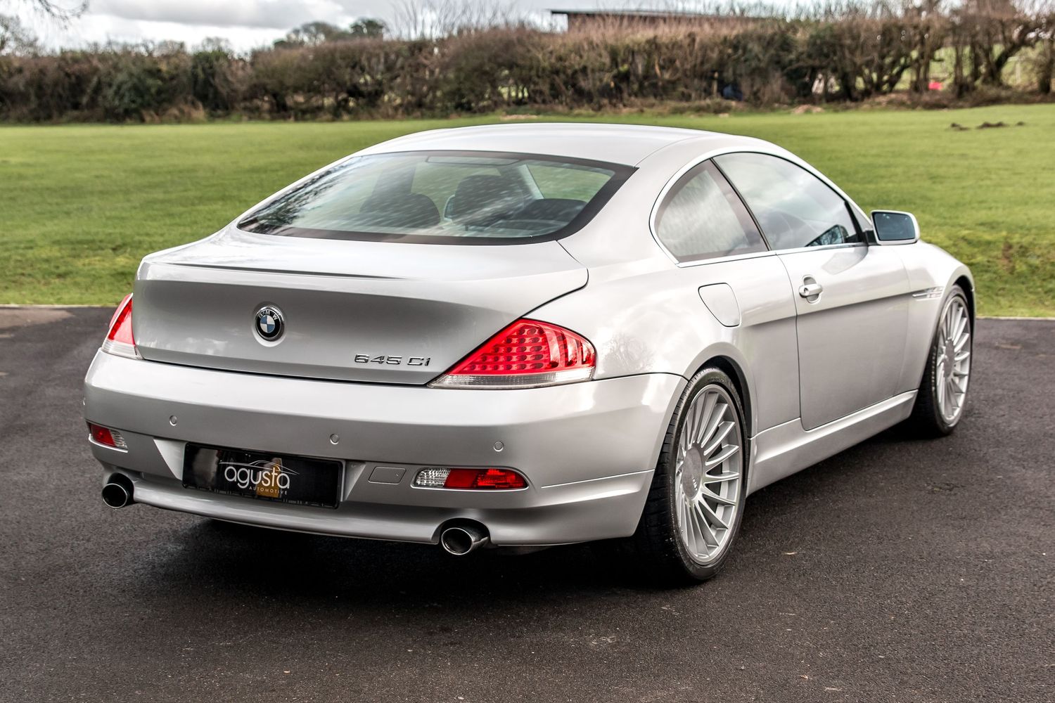 Silver BMW 6 Series coupe rear three-quarter view parked on tarmac with green trees in background.