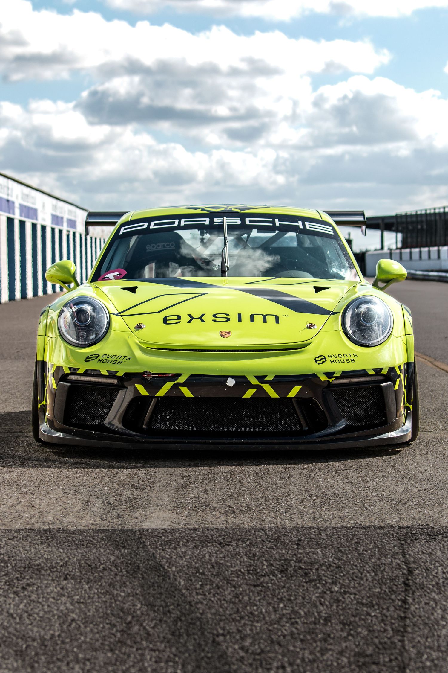 Lime green Porsche 911 GT3 race car with Exsim livery parked on a race track under cloudy skies.