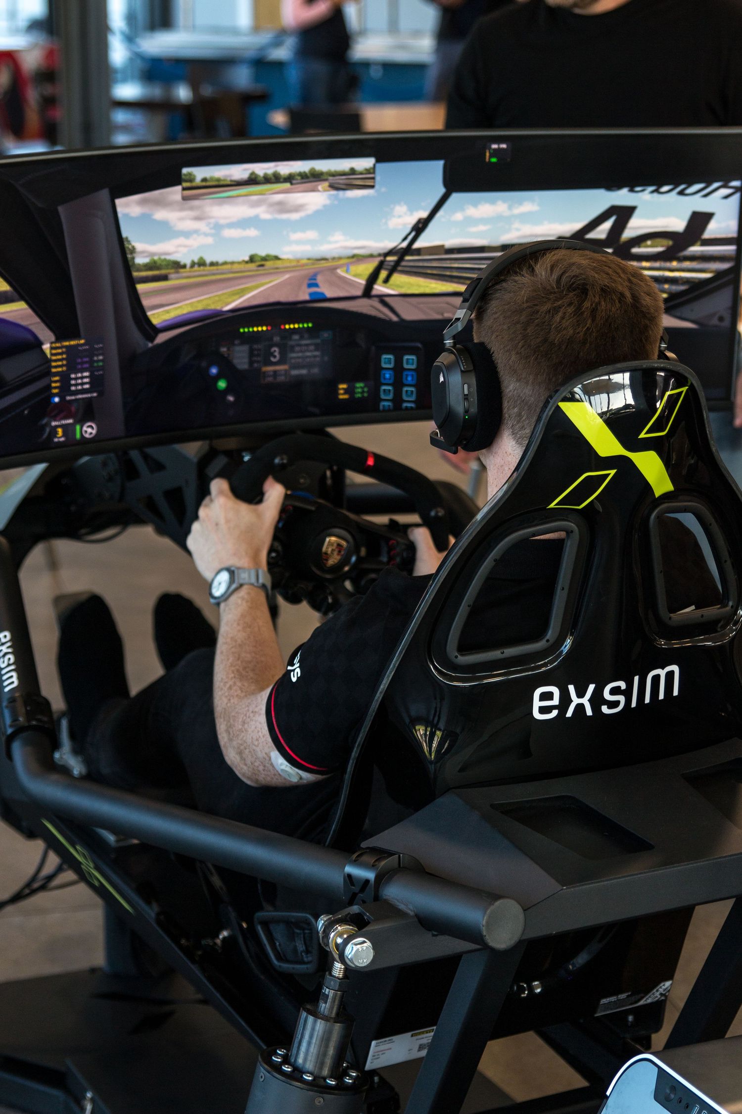 Man using an Exsim racing simulator with steering wheel and triple screens showing a race track.