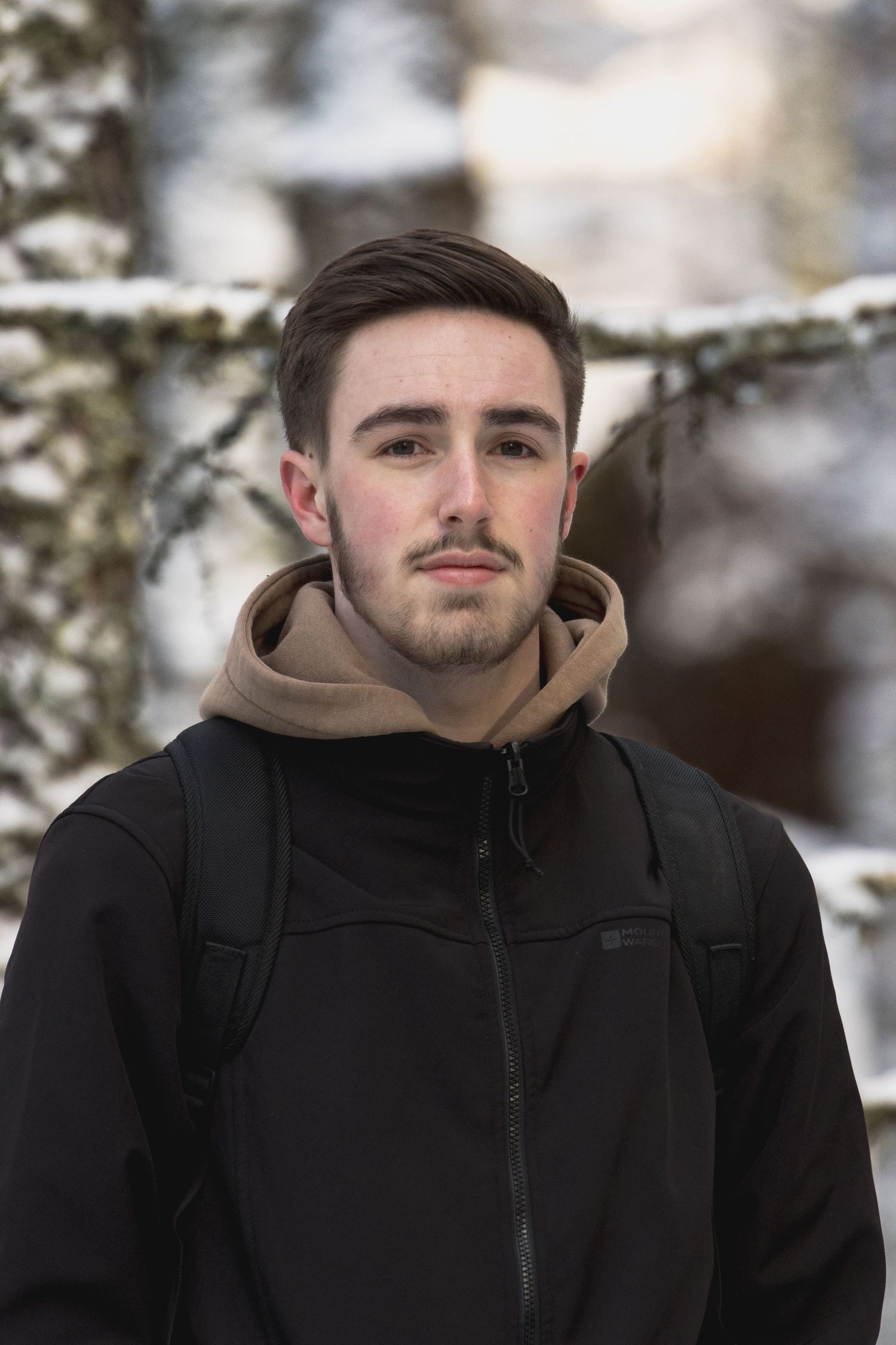 Young man with dark hair and light stubble wearing a black jacket and tan hoodie, standing in a snowy winter forest.