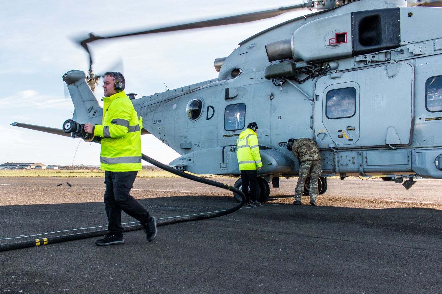 Military ground crew in high-vis jackets refueling a helicopter on an airfield tarmac.