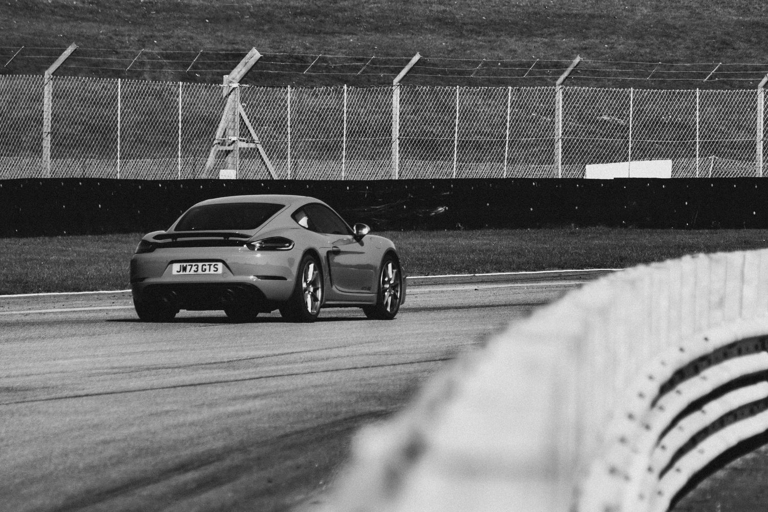 Black and white rear view of a Porsche 718 Cayman driving on a race circuit.