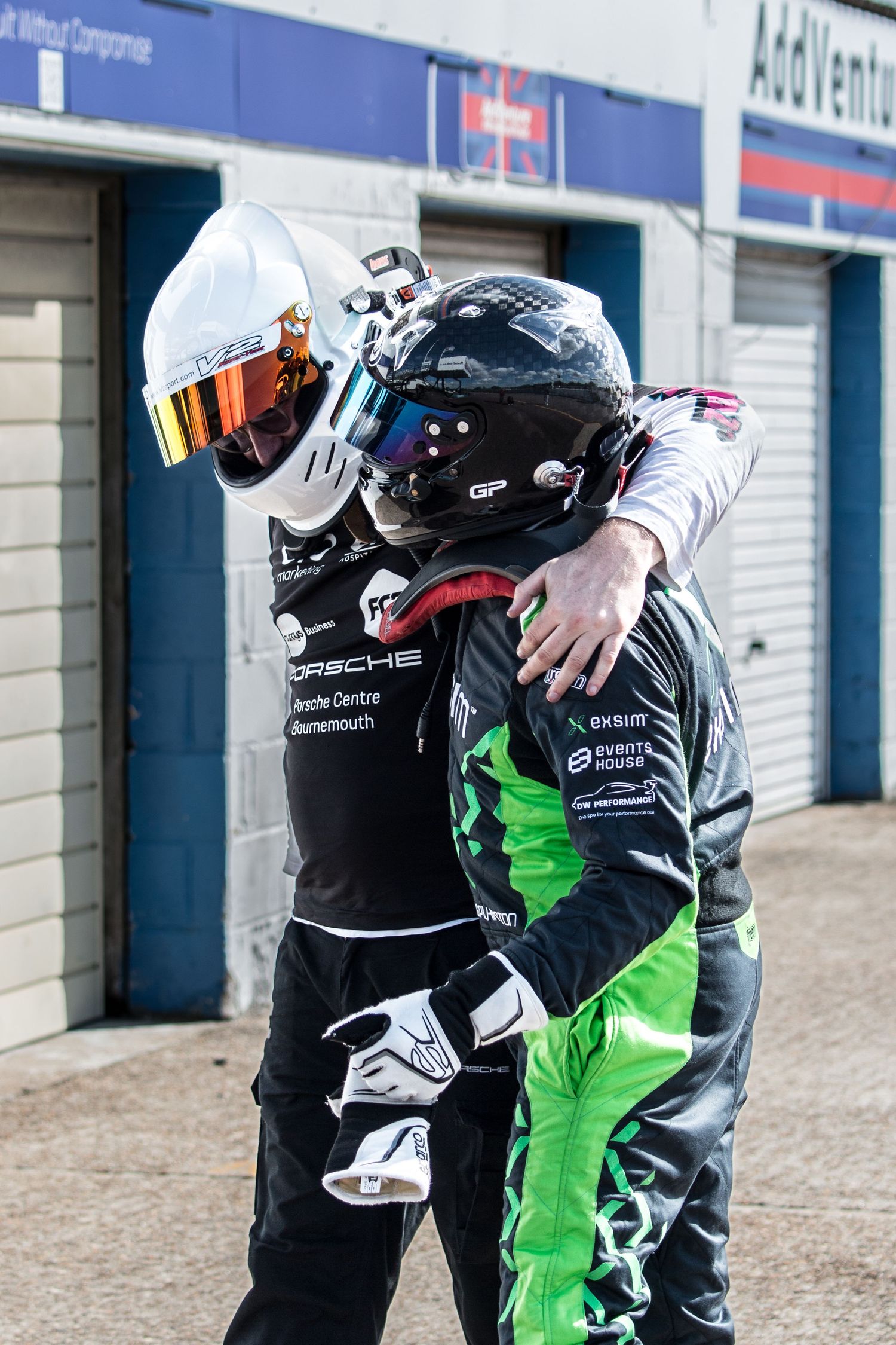 Two racing drivers in helmets and race suits embracing in the pit lane after a motorsport event.
