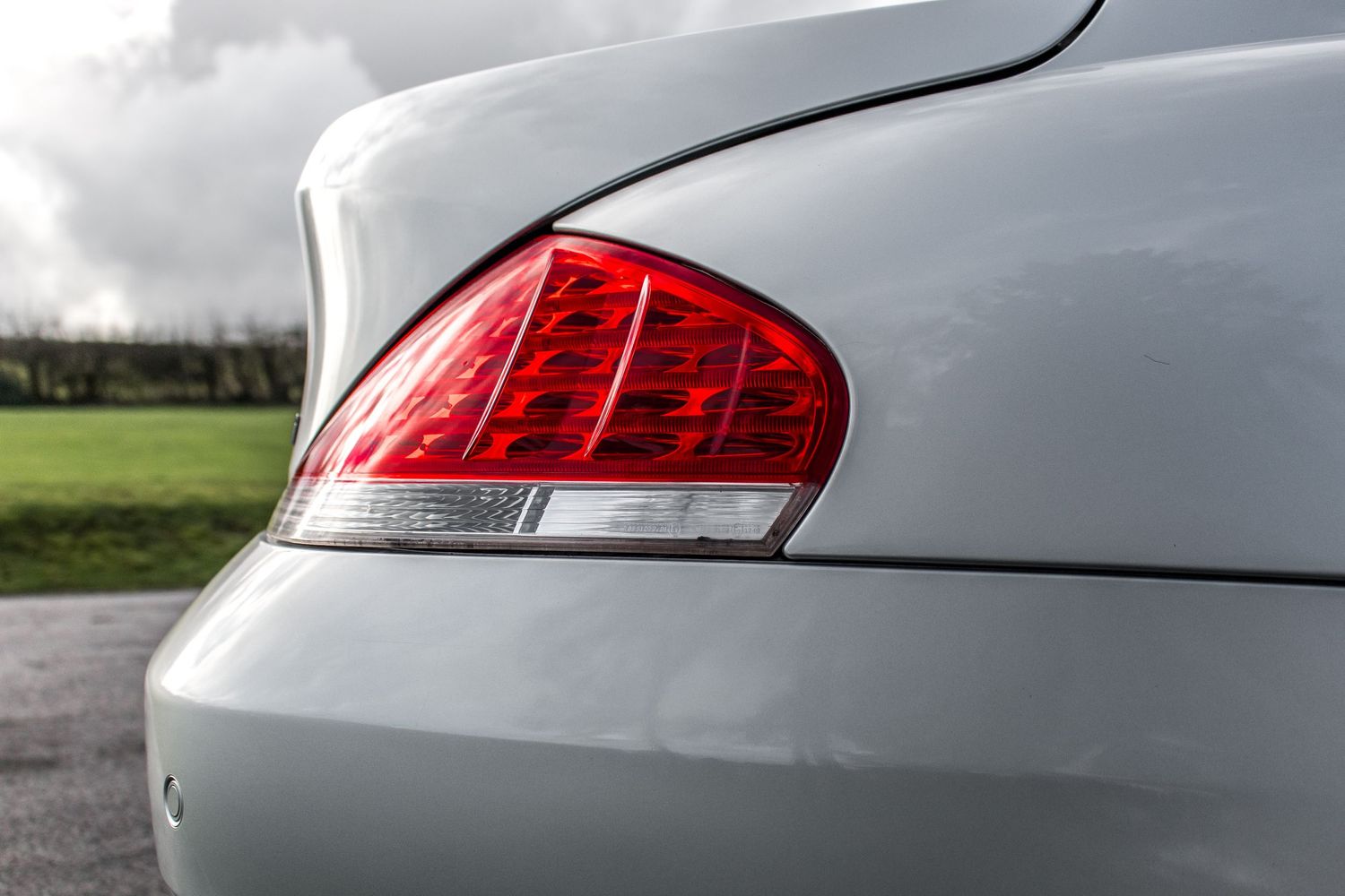 Close-up of a silver car's red tail light against a cloudy sky with green fields in the background.