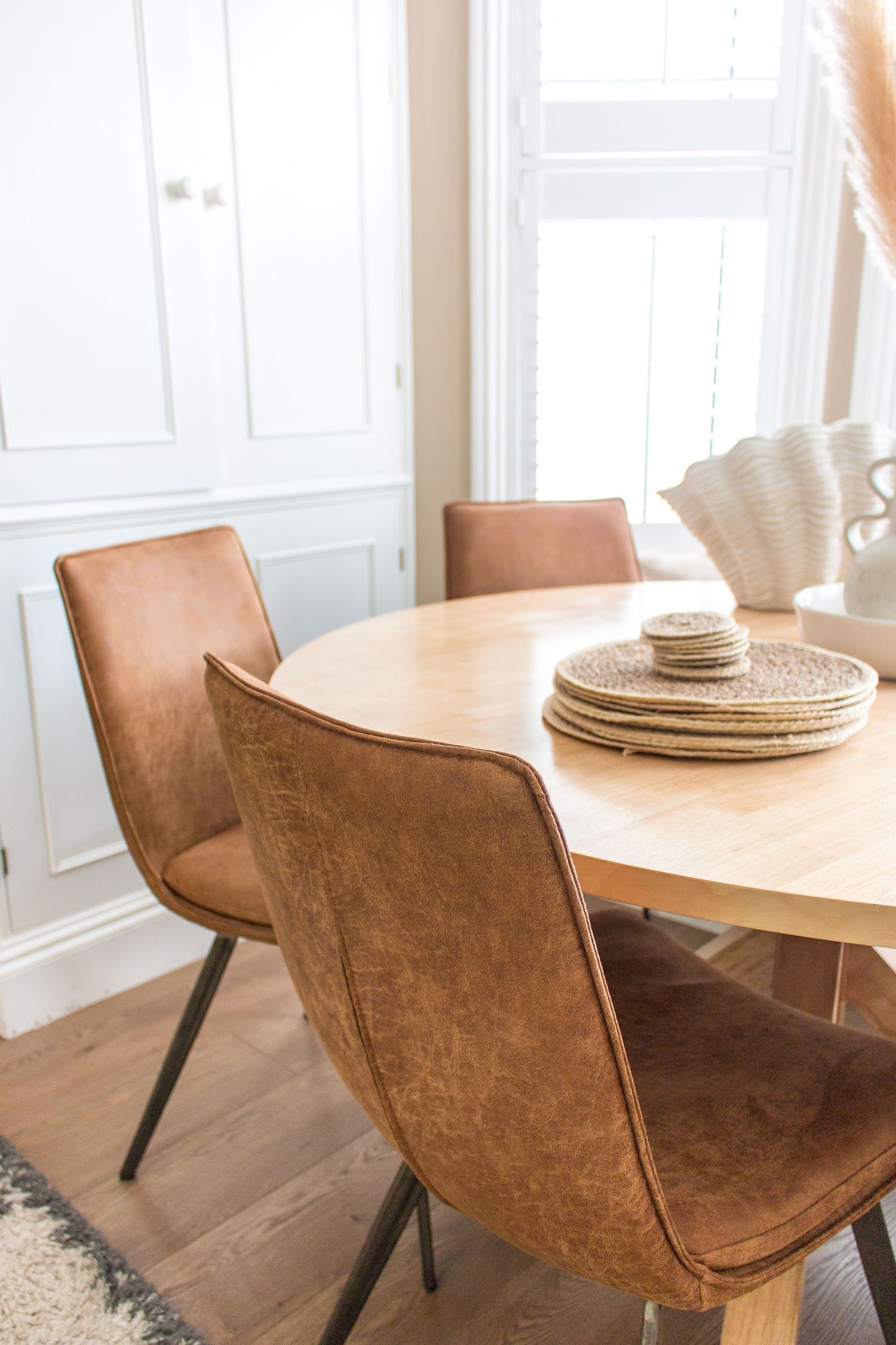 Round wooden dining table with tan leather chairs in bright white room.