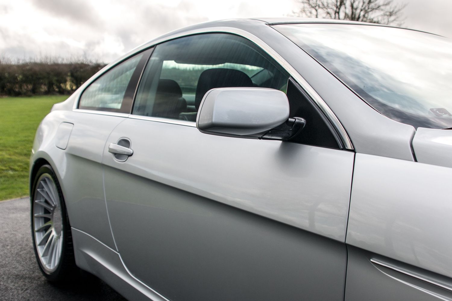 Silver BMW coupe showcasing sleek side profile with chrome mirror and alloy wheels on a green background.