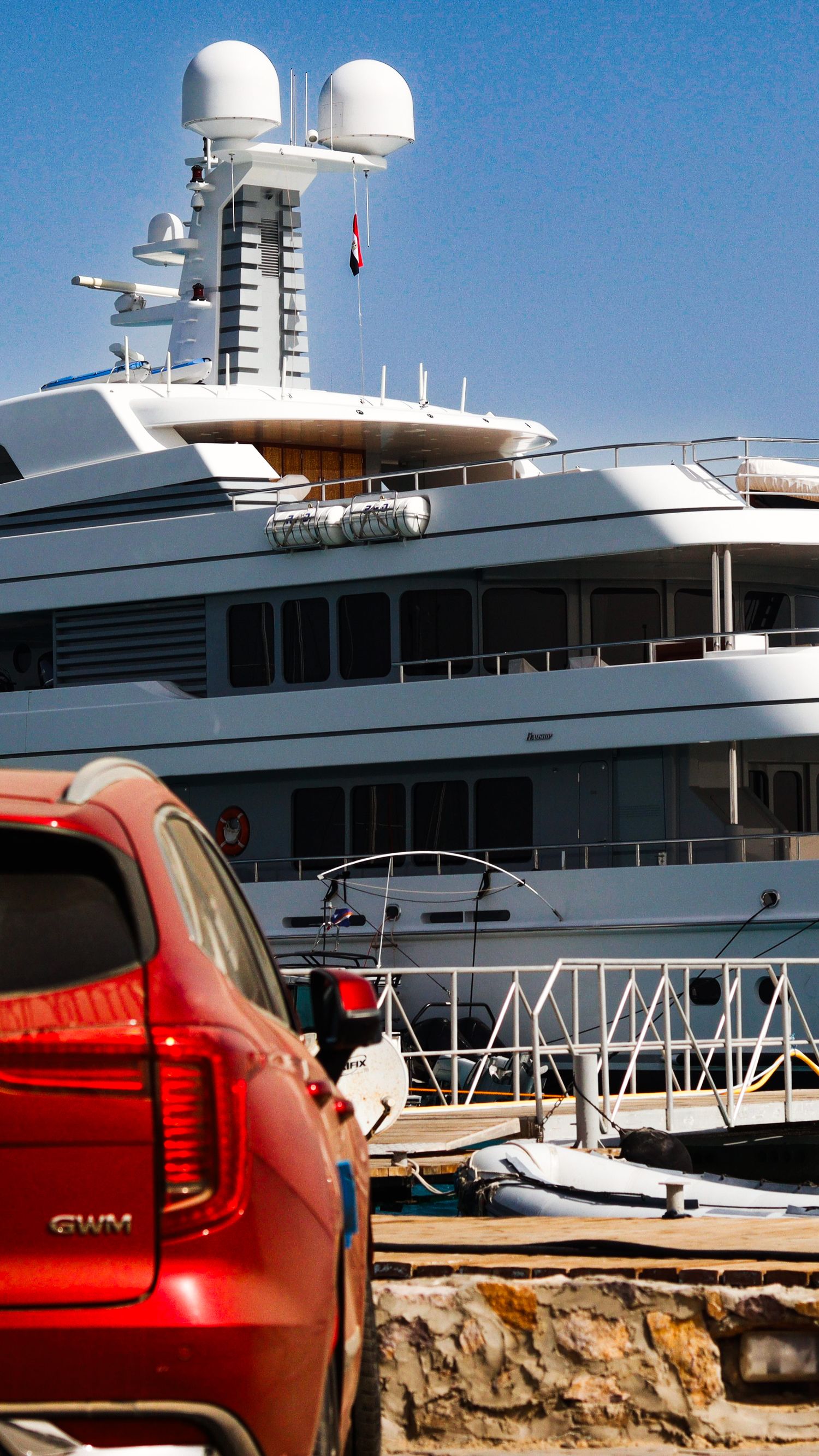 Large luxury yacht docked at a marina, with part of a red vehicle visible in the foreground under a clear blue sky.