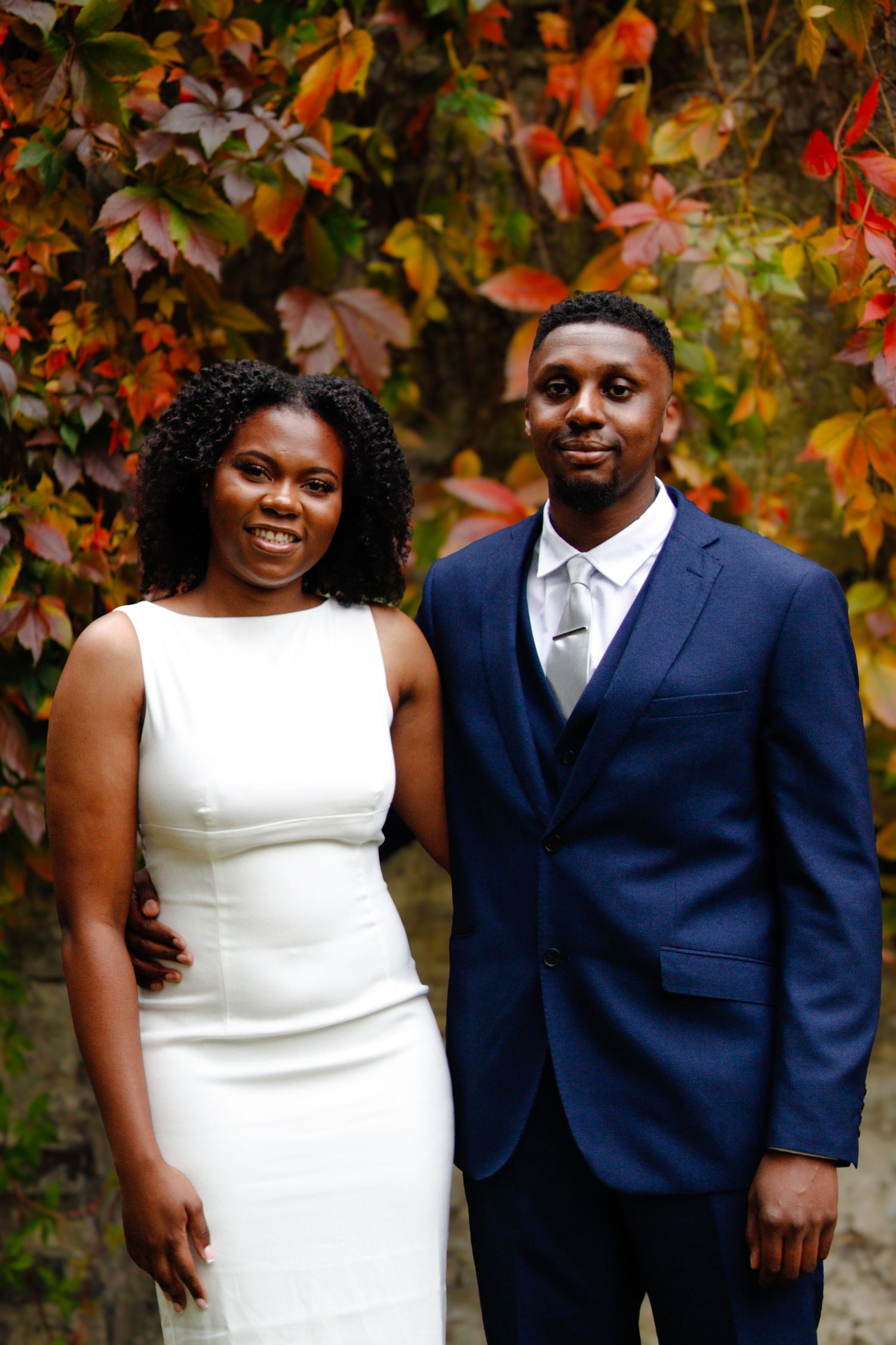 Bride and groom posing together outdoors in front of vibrant autumn foliage, with the bride in a light dress and the groom in a blue suit.