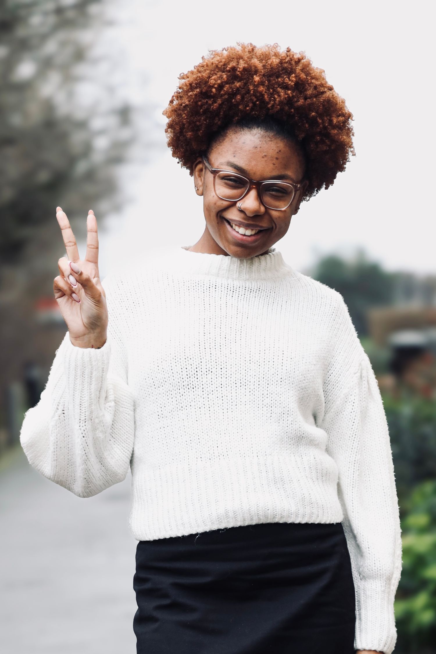 Joyful outdoor portrait of a person in a cozy white sweater smiling and flashing a peace sign in natural light.