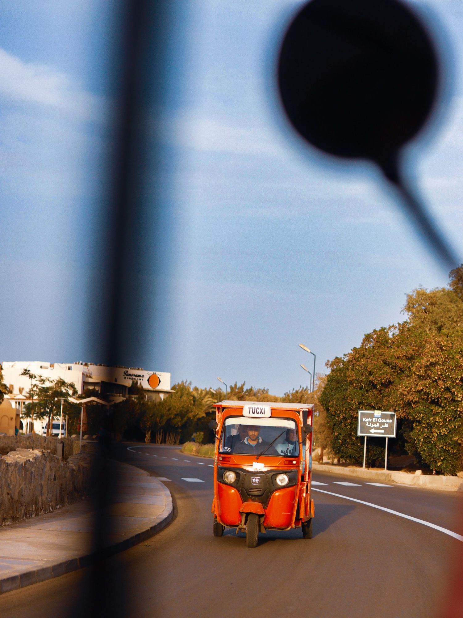 Street scene photographed from inside a moving vehicle, with a red tuk-tuk driving along a sunlit road.