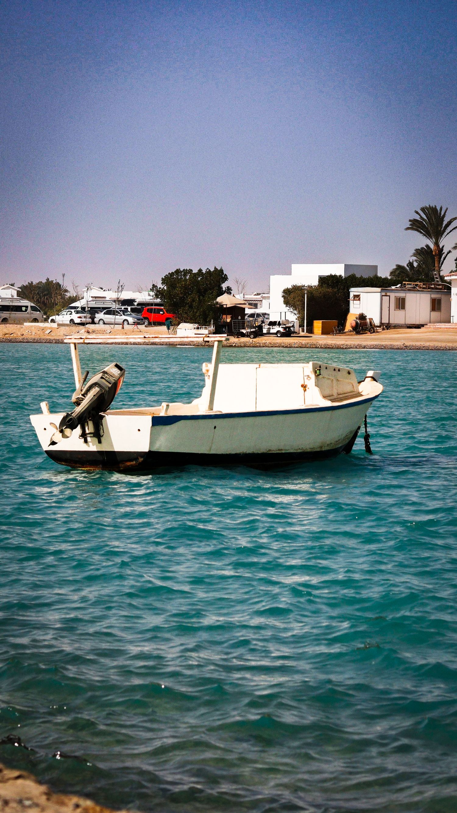 Small white boat floating on clear turquoise water near a sunny shoreline with buildings in the background.