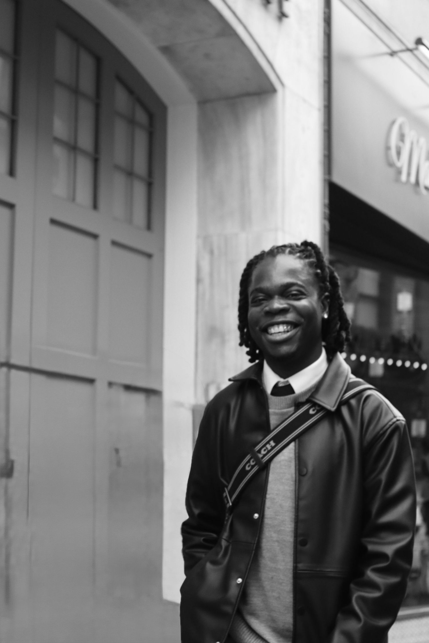 Black and white portrait of a smiling man in a leather jacket standing near a city building, capturing a candid street moment.