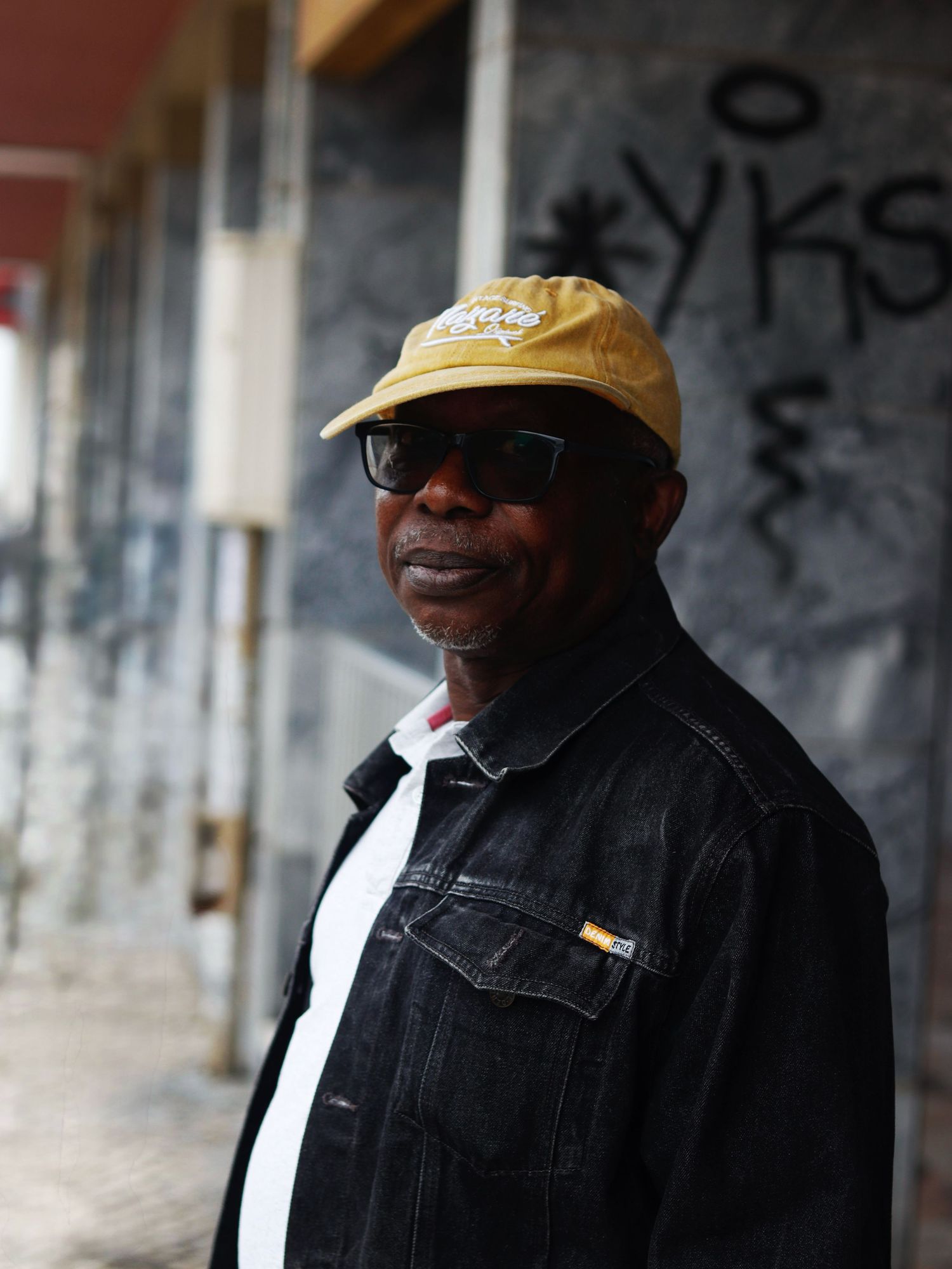 Stylish portrait of a man in a yellow cap and dark jacket standing against a textured urban wall with graffiti.