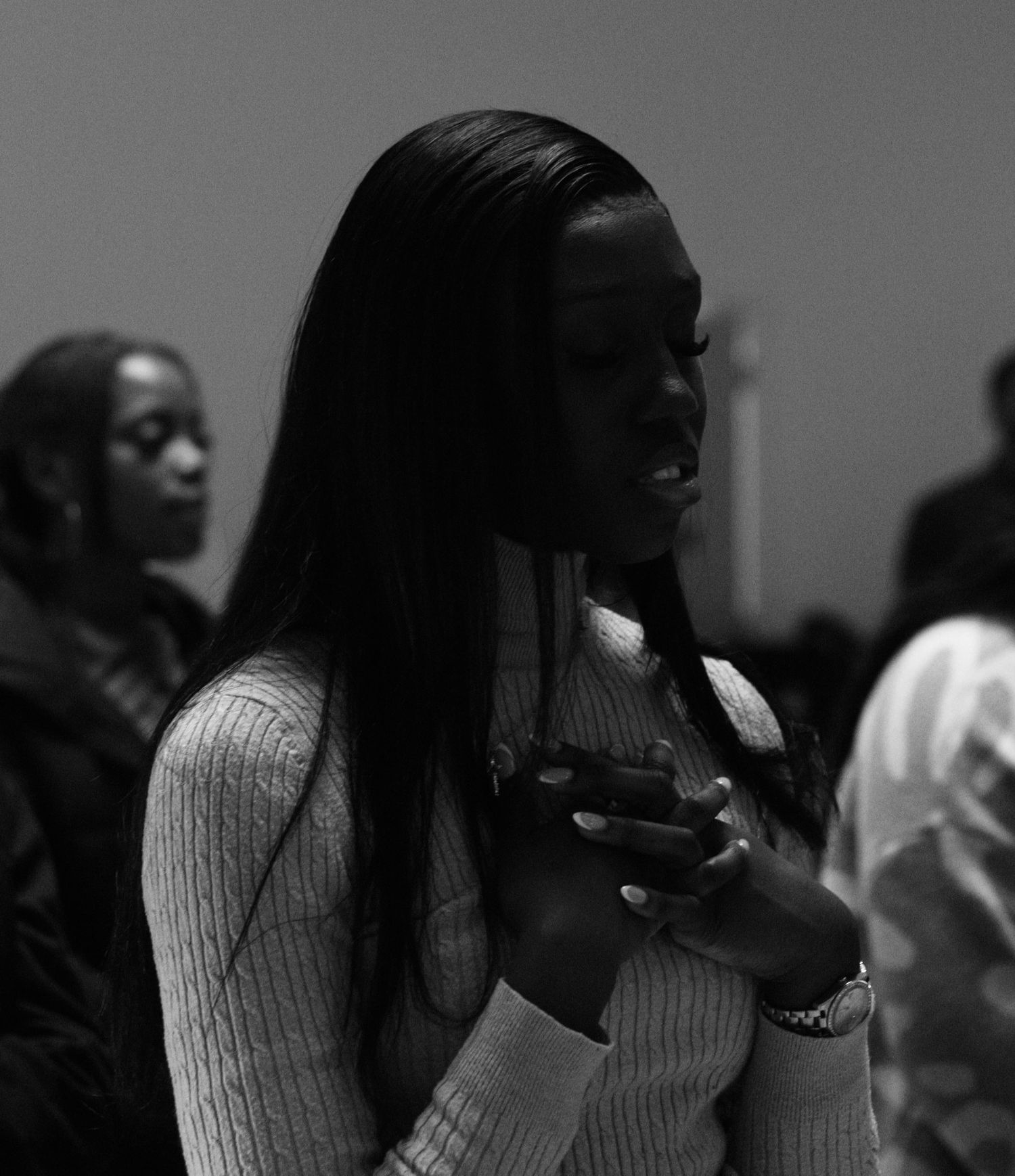 Black and white photo of a woman in a church service, standing with hand over her chest in a reflective worship moment.