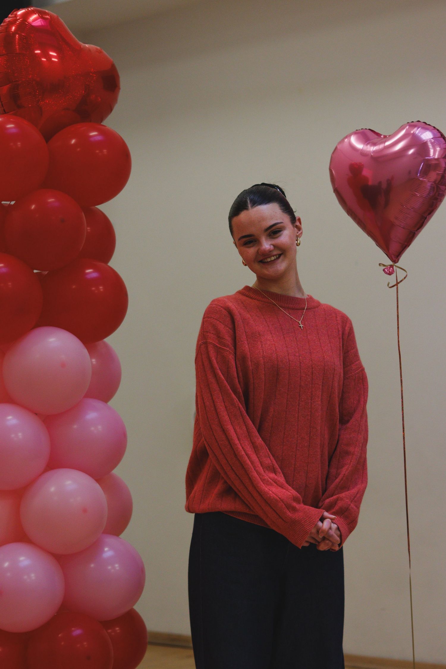 Person in a red sweater standing beside a display of red and pink balloons, holding a heart-shaped balloon against a neutral background.