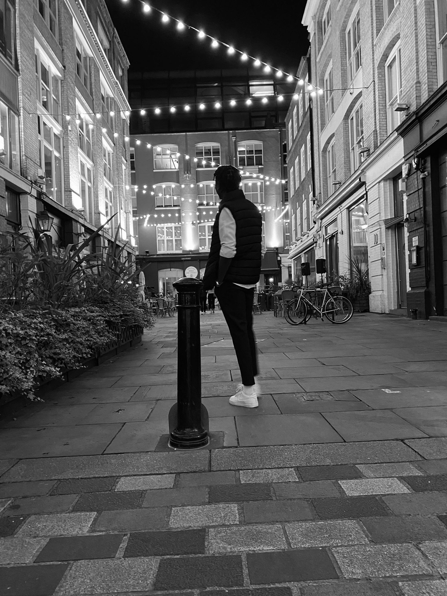 Person walking down a city street at night, captured in black and white with overhead string lights creating a dramatic urban scene.
