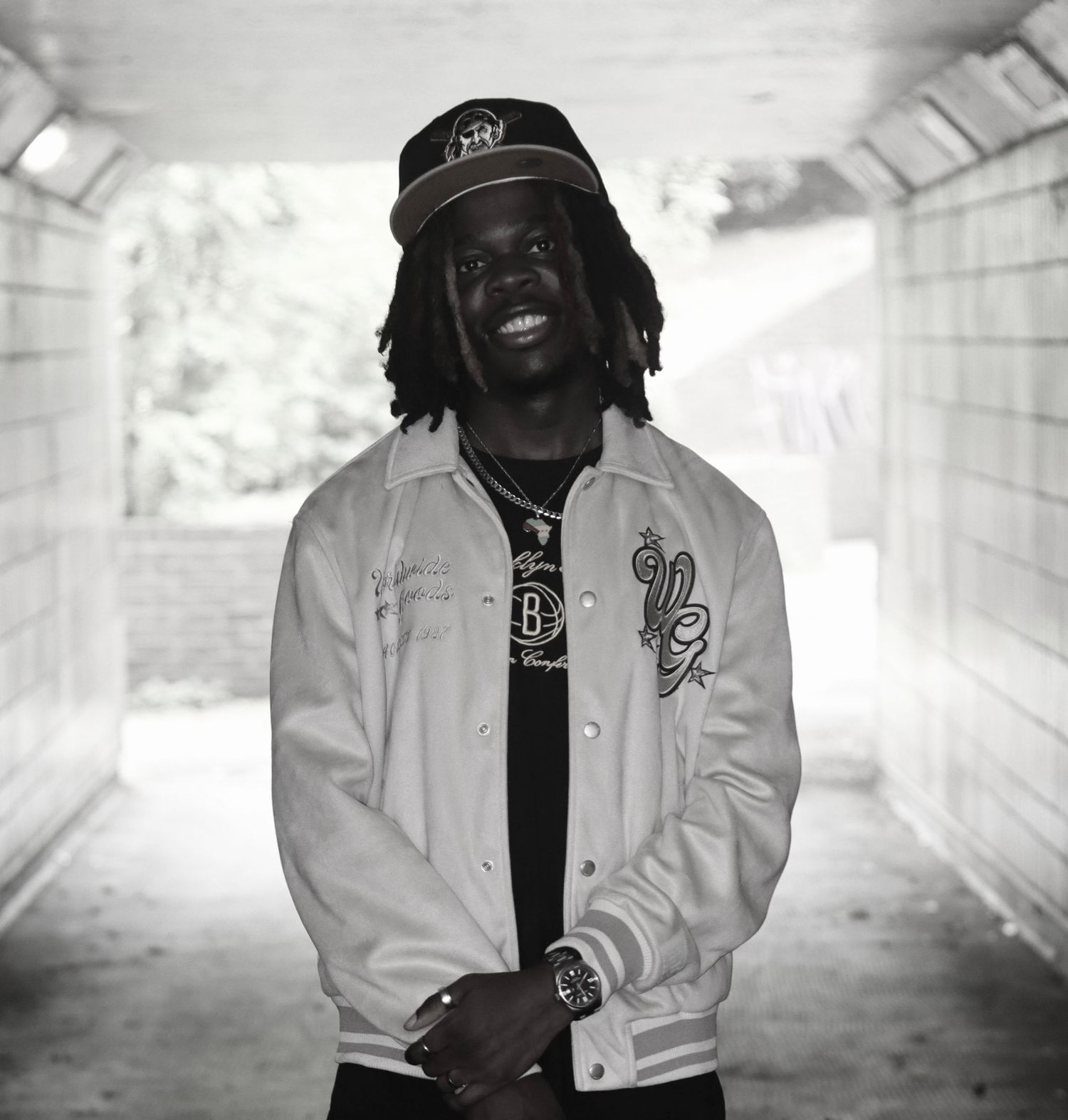 Black and white portrait of a man standing in a softly lit tunnel, wearing casual streetwear and looking confidently at the camera.