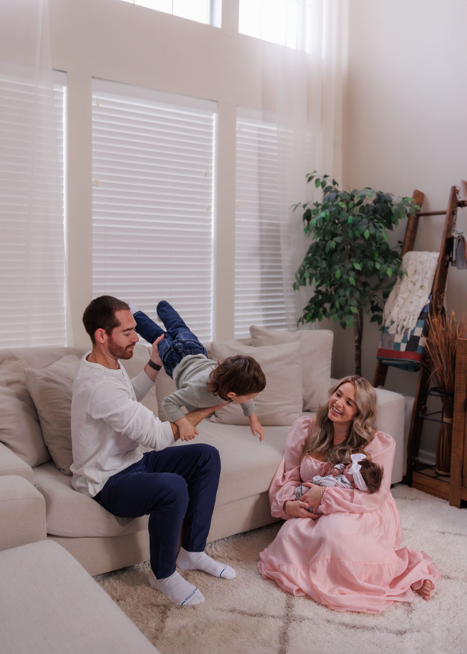 family of 4 in living room, dads lifting young child in air while mom holds newborn