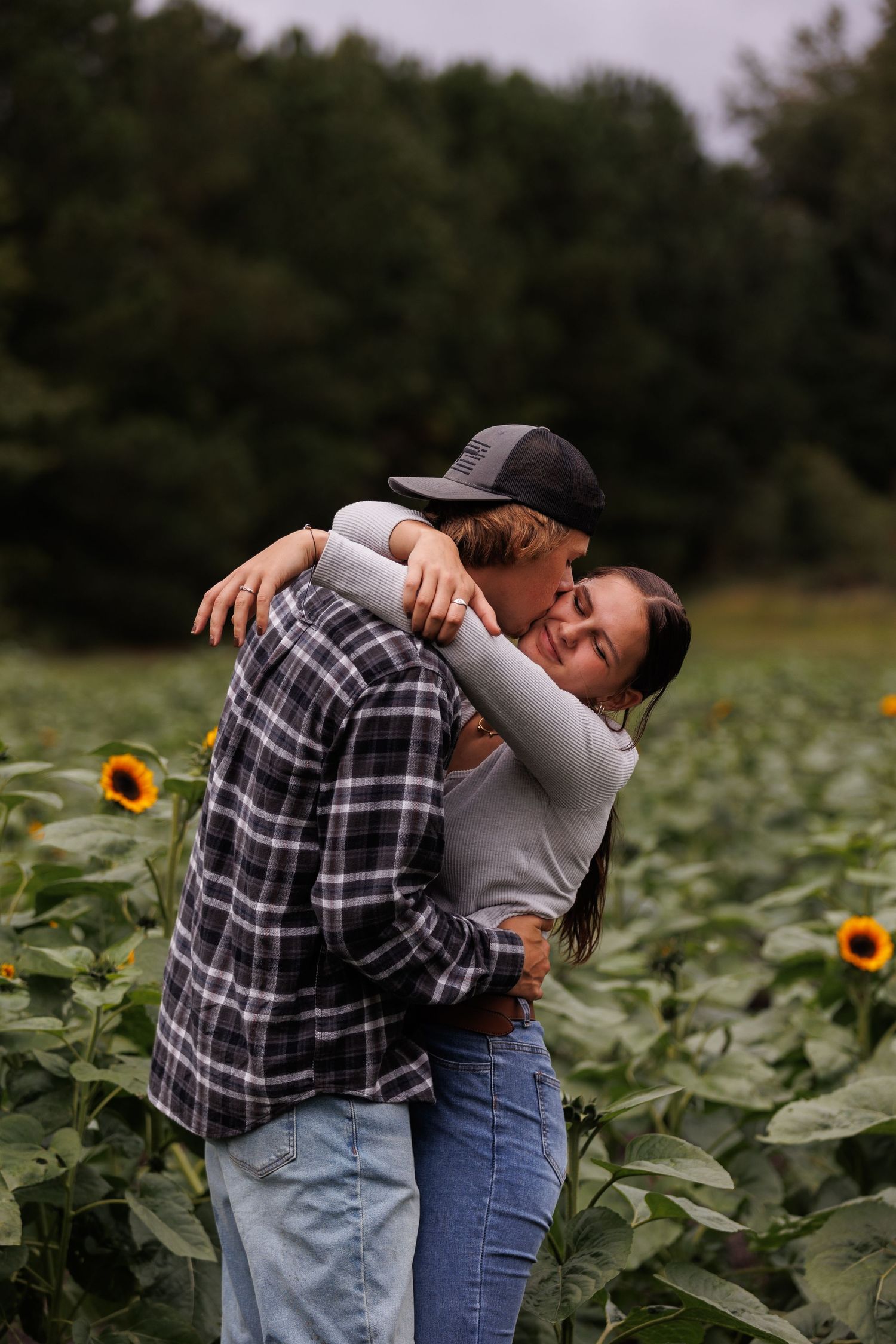 couple in sunflower field, her arms around his neck, hes kissing her cheek
