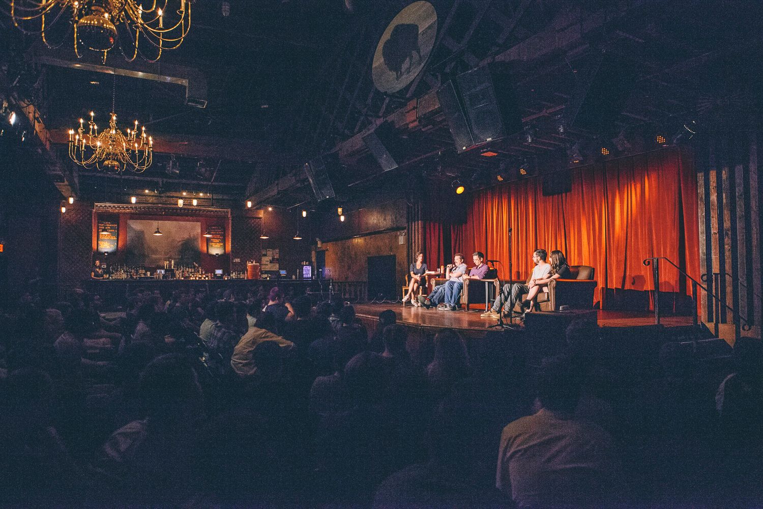 Wide shot of packed theater audience watching panel discussion on stage with red curtains.