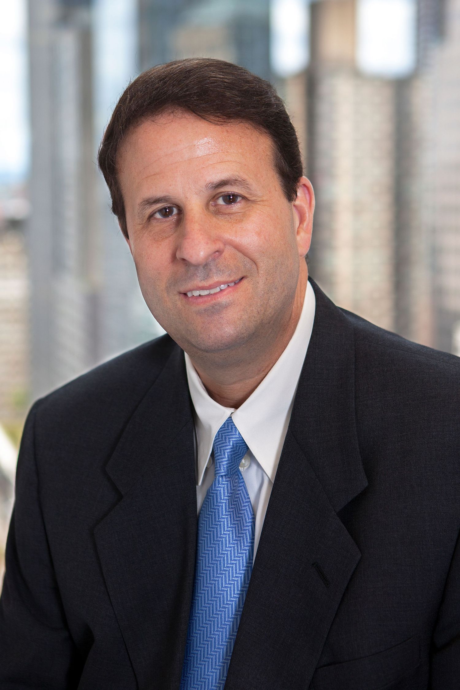 Professional man in dark suit and blue tie smiling in a modern office setting.