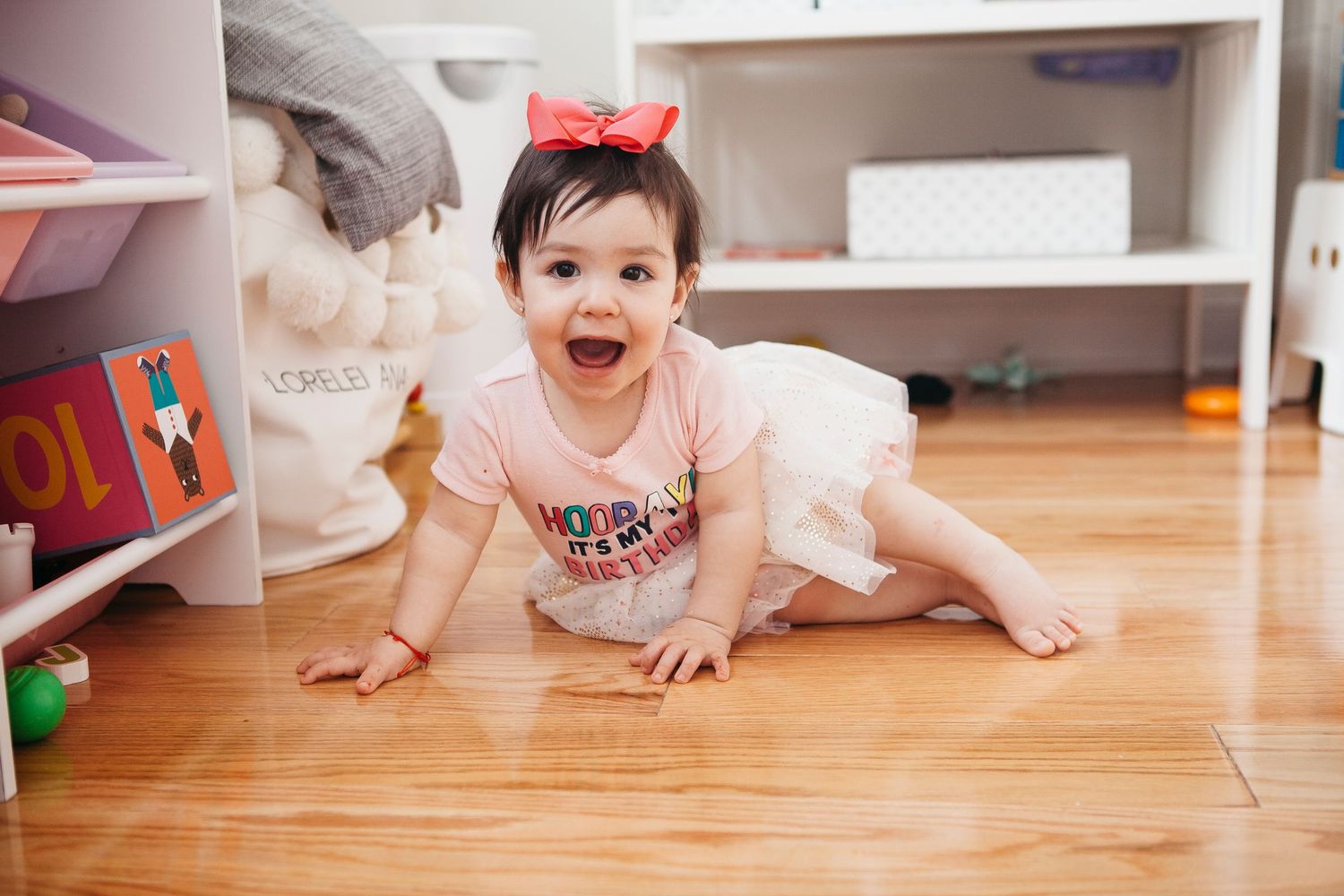 Happy baby girl in red bow and birthday shirt crawls on hardwood floor in playroom.
