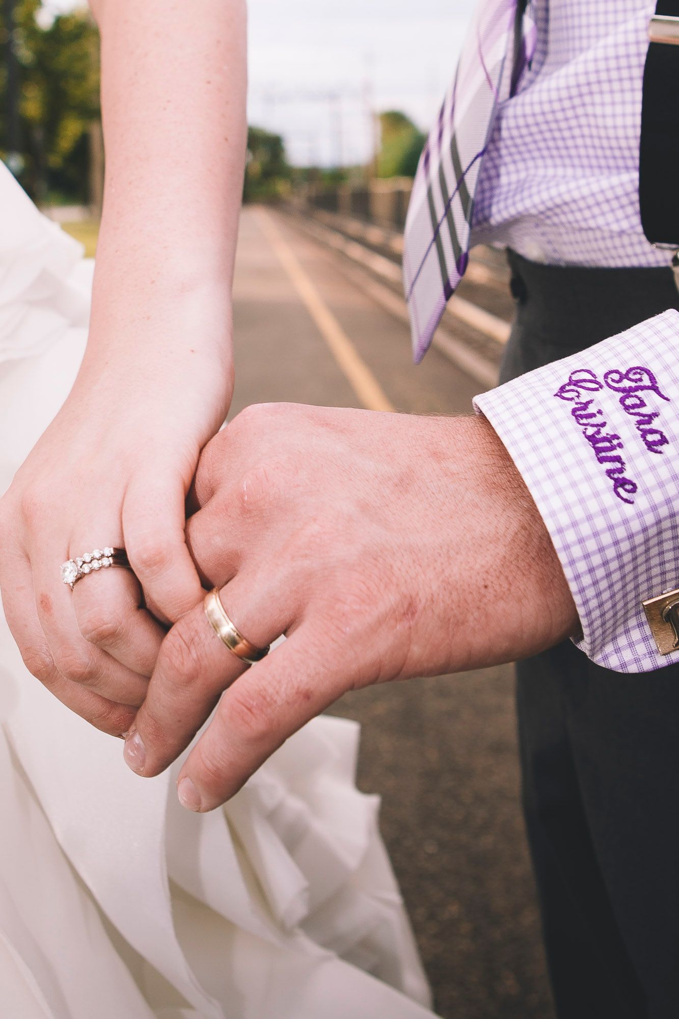 Close-up of bride and groom holding hands, showing wedding rings and embroidered shirt cuff on road.