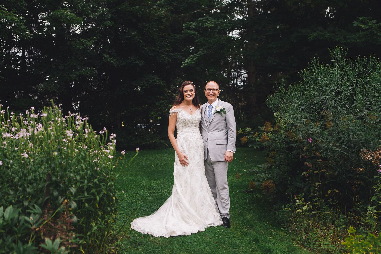Bride in lace gown and groom in grey suit posing in lush garden wedding portrait.