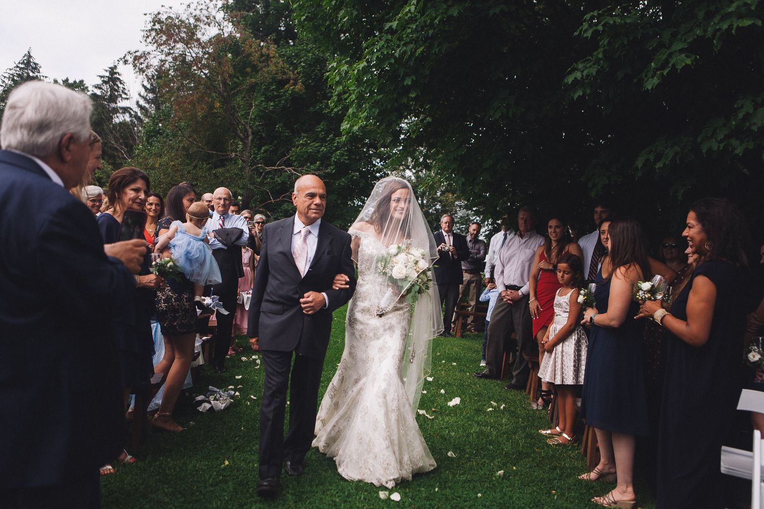 Bride in lace gown walking down outdoor aisle with escort while guests celebrate on both sides.