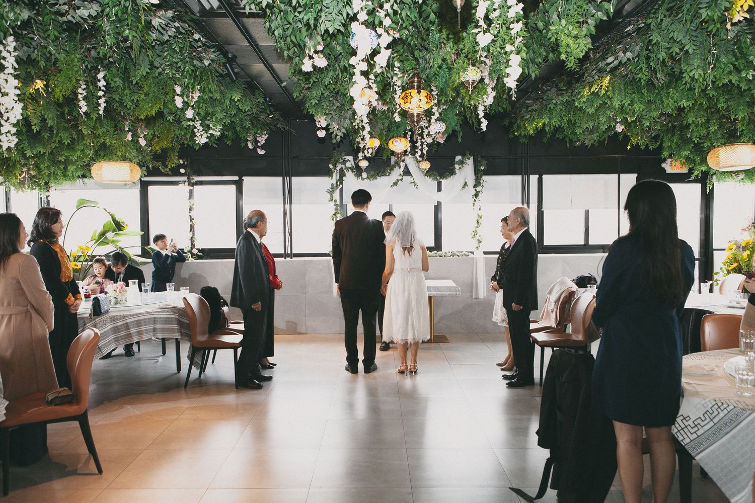 Wedding ceremony in a botanical venue with lush hanging greenery and guests lining the aisle.