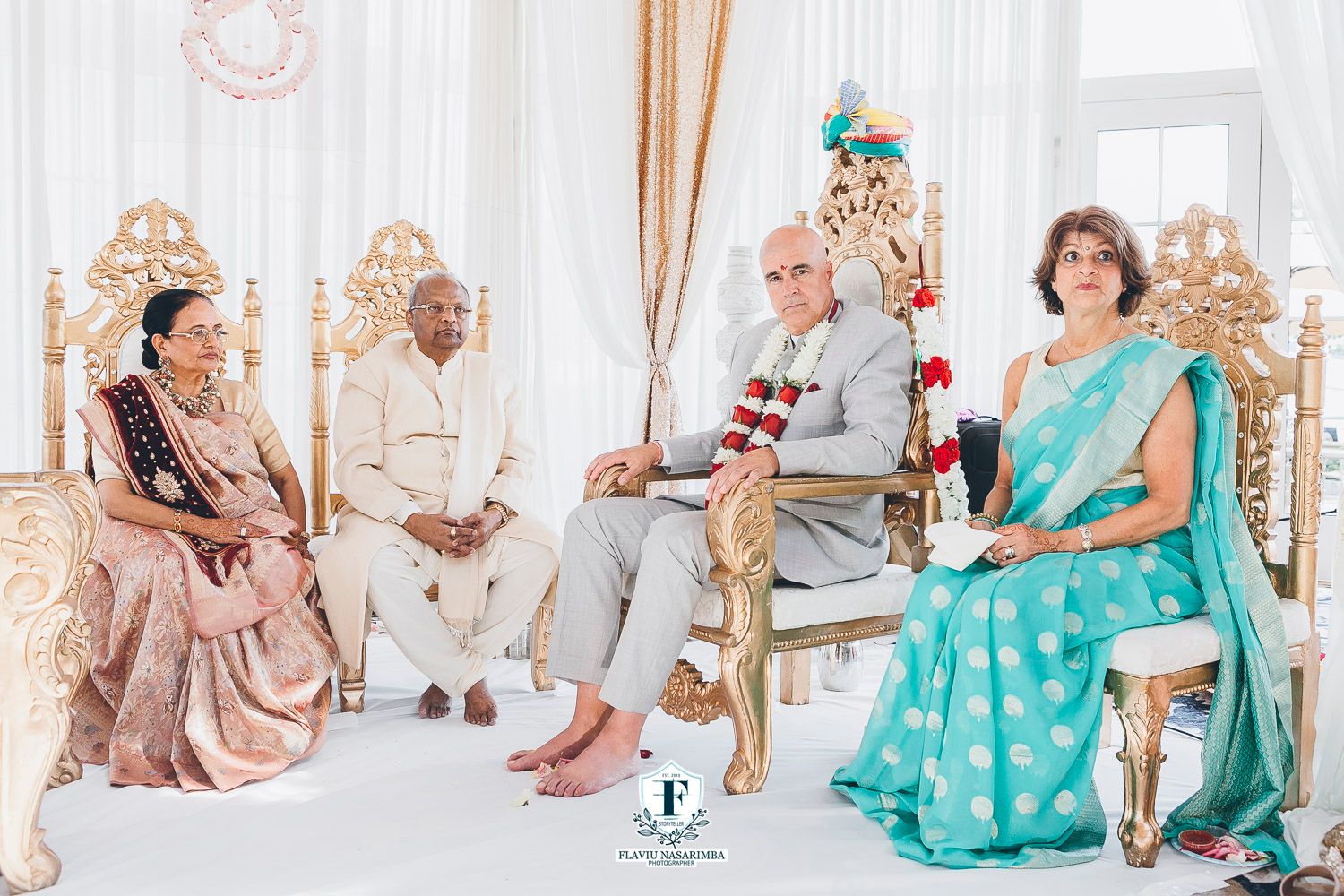 Four family members in traditional Indian attire seated on ornate gold chairs during Hindu wedding ceremony.
