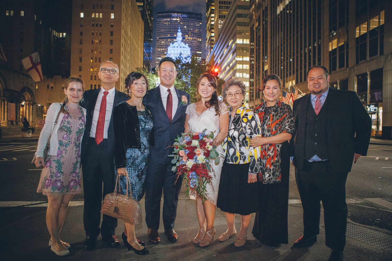 Wedding party group photo on city street at night with colorful bouquet and Empire State Building lit up.