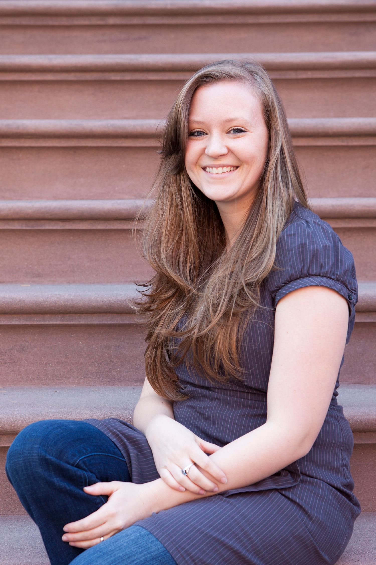 Young woman with long brown hair smiling, wearing a gray dress and jeans, sitting on steps.