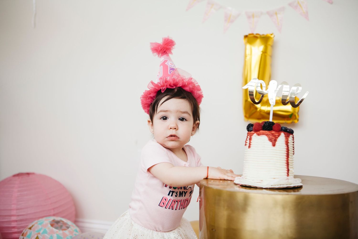 Baby girl in pink party hat stands at gold pedestal table beside her decorated first birthday cake.