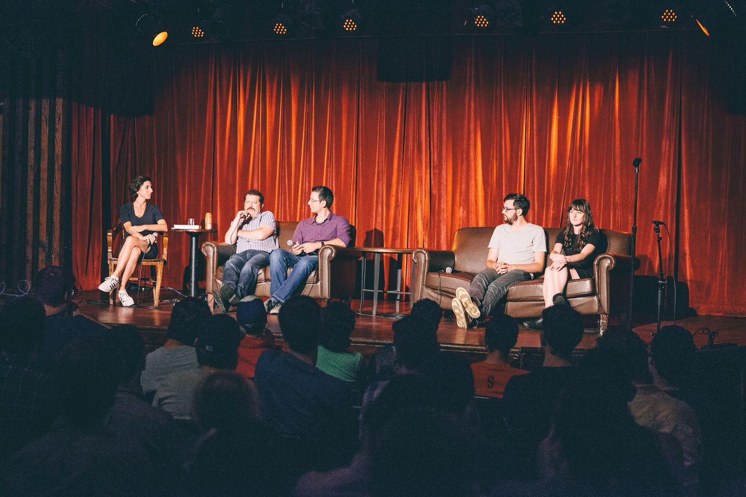 Five panelists seated in chairs on stage during discussion event with red curtain backdrop.