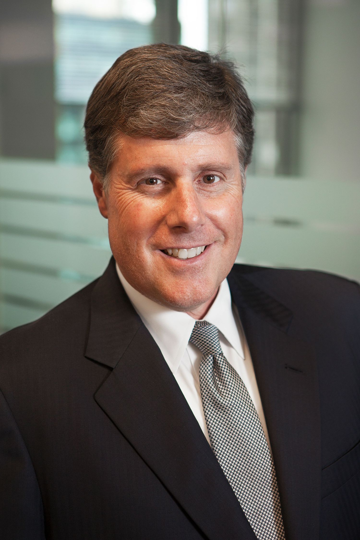 Professional man in dark suit and gray tie smiling in a modern office setting.