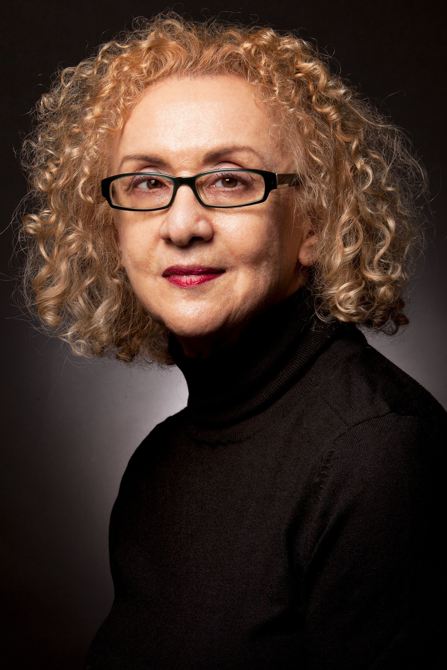 Older woman with curly blonde hair and black glasses wearing a black turtleneck, studio portrait.