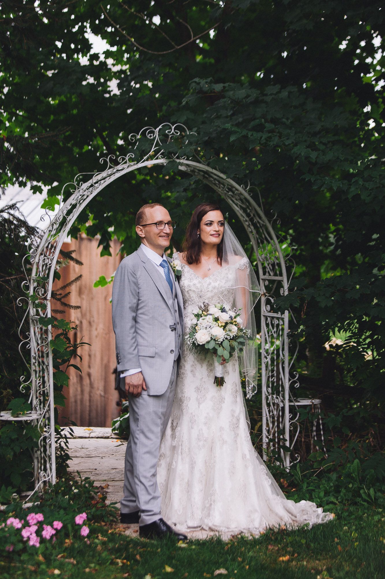 Bride and groom posing under a decorative white iron arch surrounded by green foliage and flowers.
