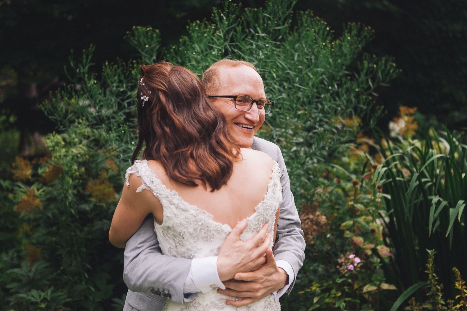 Groom embracing bride in lace backless wedding gown in romantic garden portrait.