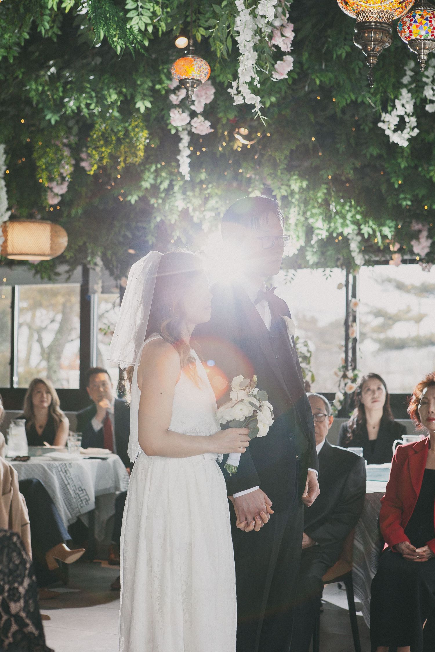 Bride and groom share first kiss under lush green canopy with sunlight flare at ceremony.
