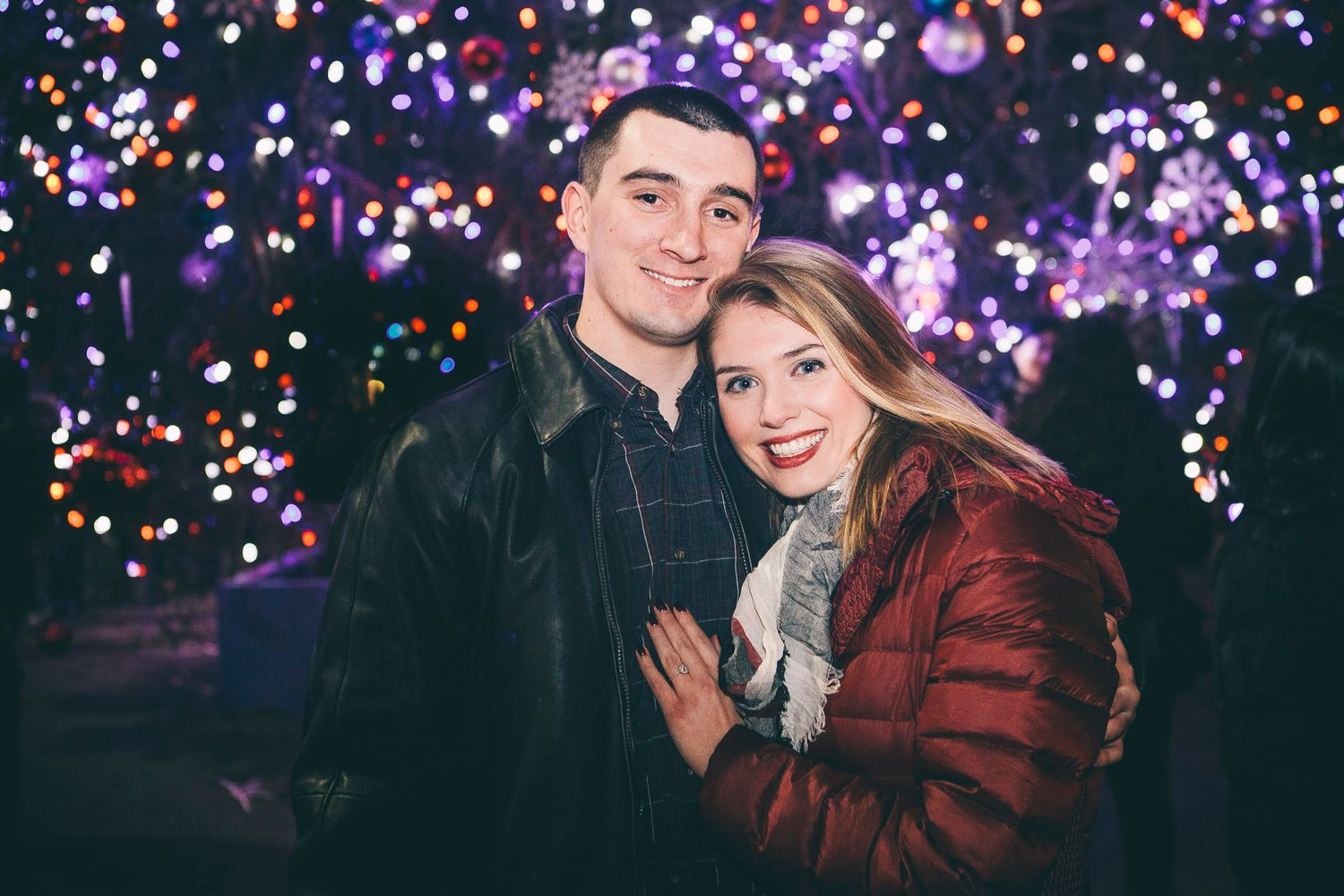Smiling couple embraces before bokeh Christmas tree lights in colorful nighttime holiday setting.
