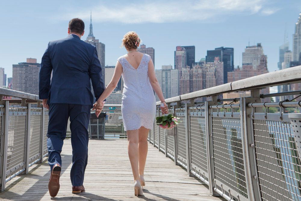 Bride and groom walk hand in hand on waterfront boardwalk with NYC skyline in the background.