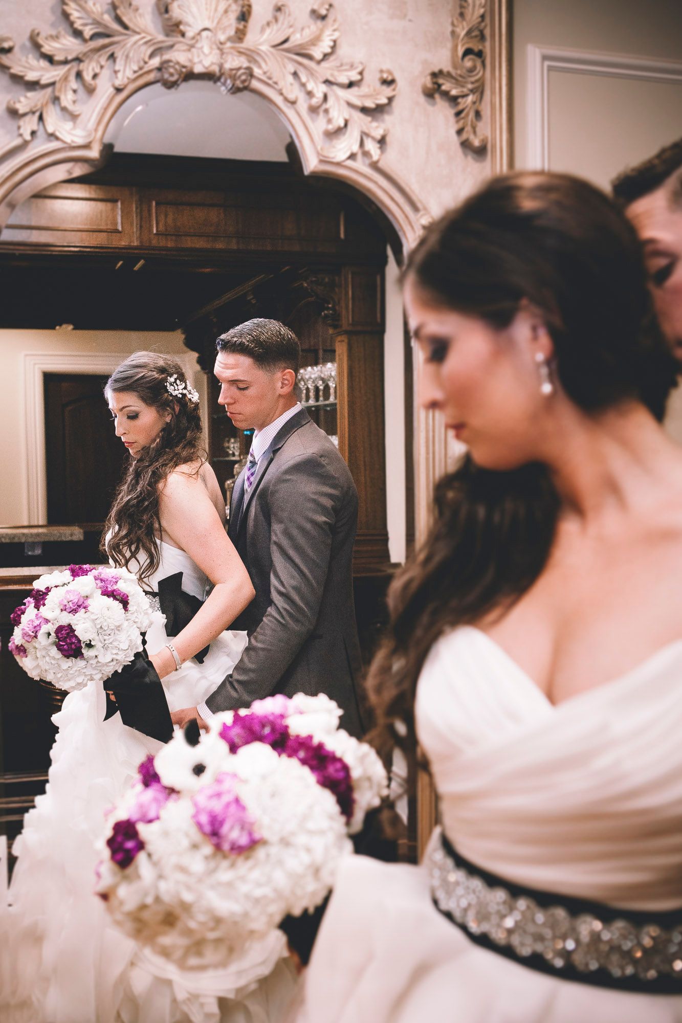 Bride and groom walk with bridesmaids holding purple and white bouquets in an ornate venue.