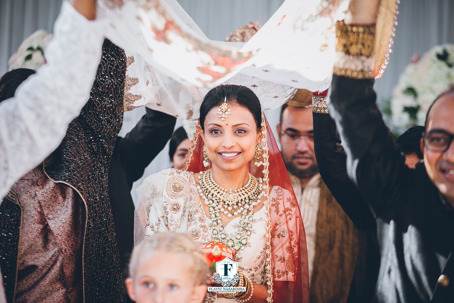 Smiling Indian bride in white and gold lehenga with traditional jewelry during wedding ceremony procession.