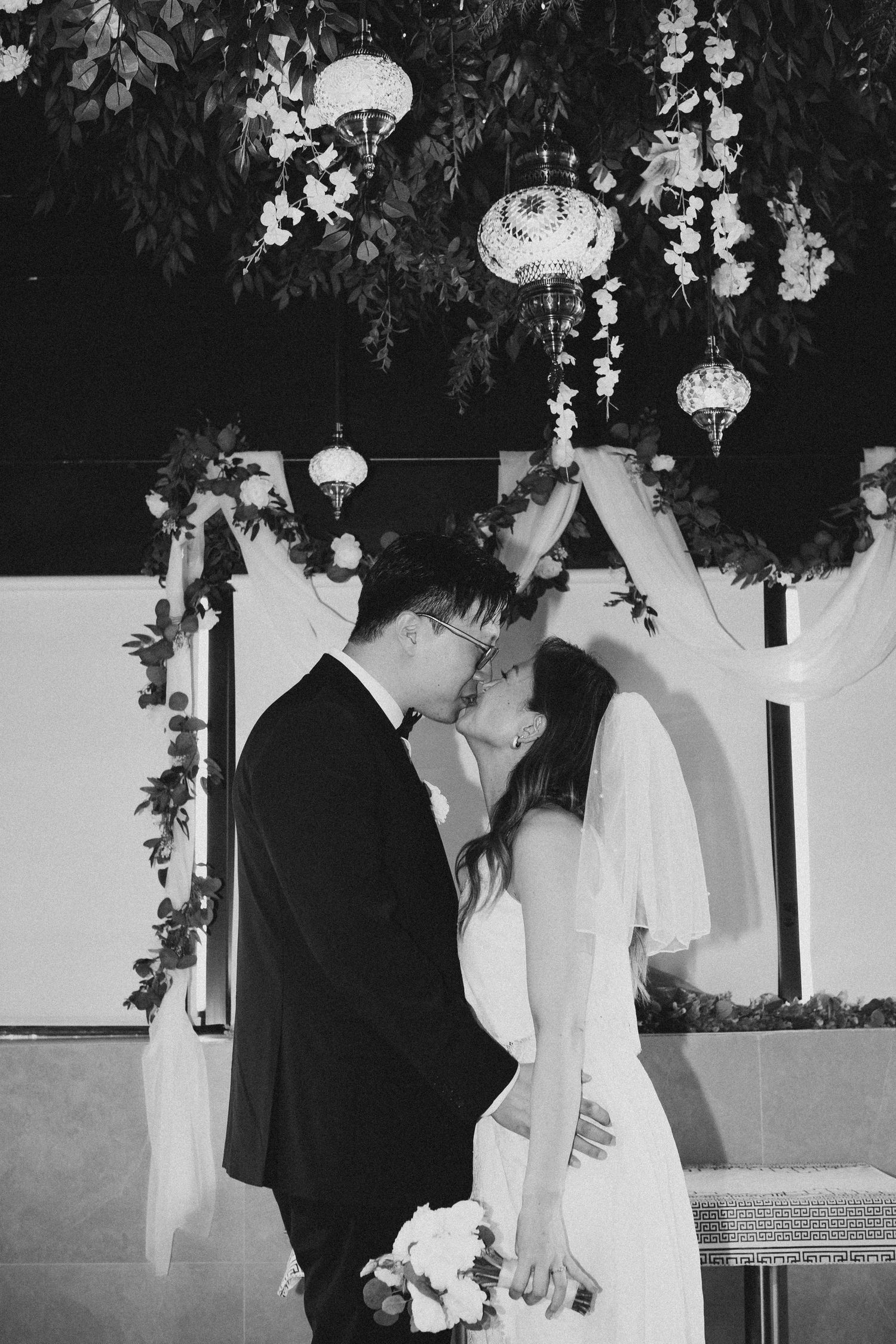 Black and white photo of bride and groom kissing under floral ceremony arch.