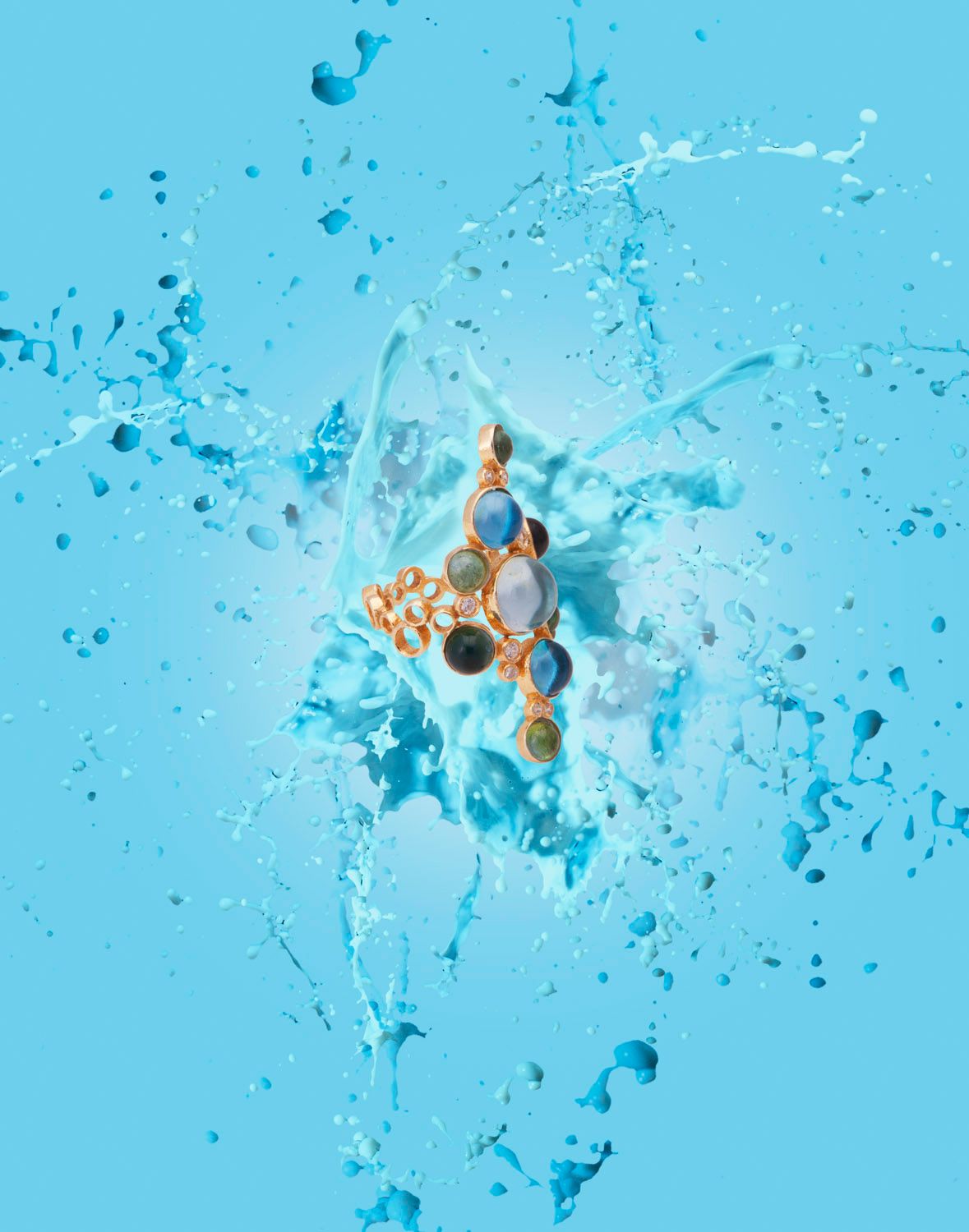 Gold ring with blue and teal gemstones surrounded by water splash on blue background.