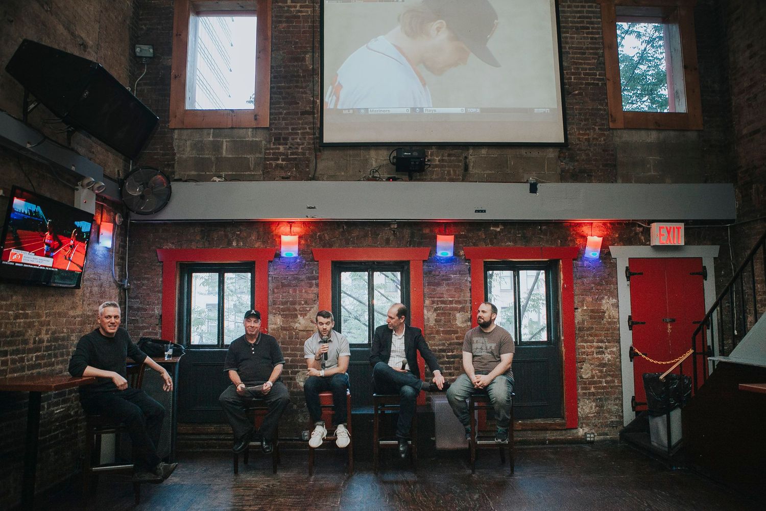 Five men seated on stools in a brick-walled venue with red doors and a projection screen behind them.